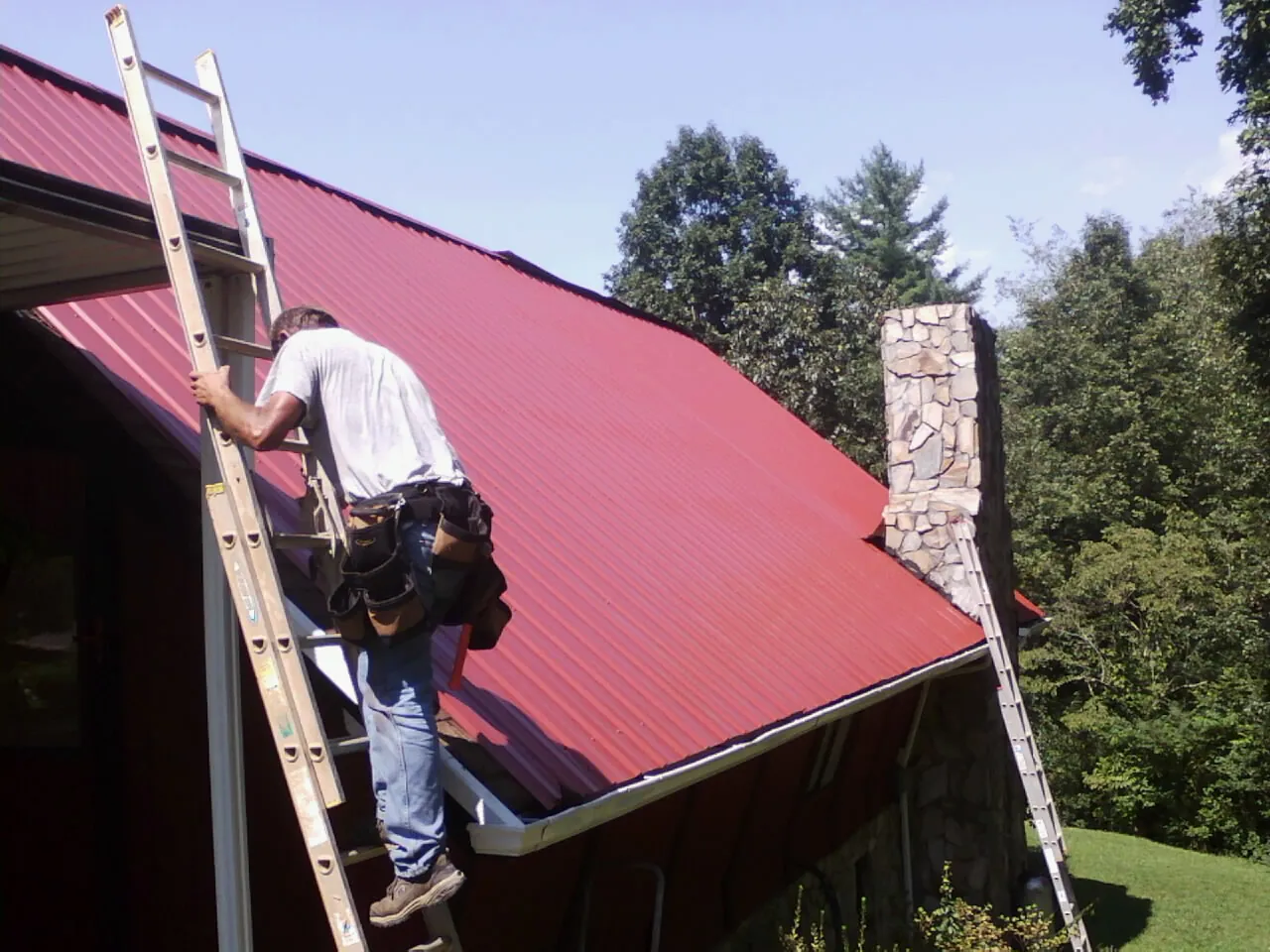 Roofer on a ladder beside a red metal roof and stone chimney — Soffit & Fascia Repair by Roofing Brothers in Camden